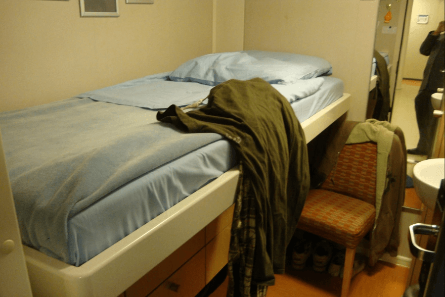 A small cruise ship cabin featuring a single elevated bed with light blue bedding and a slightly wrinkled blanket. A chair with a patterned cushion sits next to the bed, with clothes draped over it. A narrow mirror reflects part of the room, showing a person taking the photo, while a small sink is visible to the right.