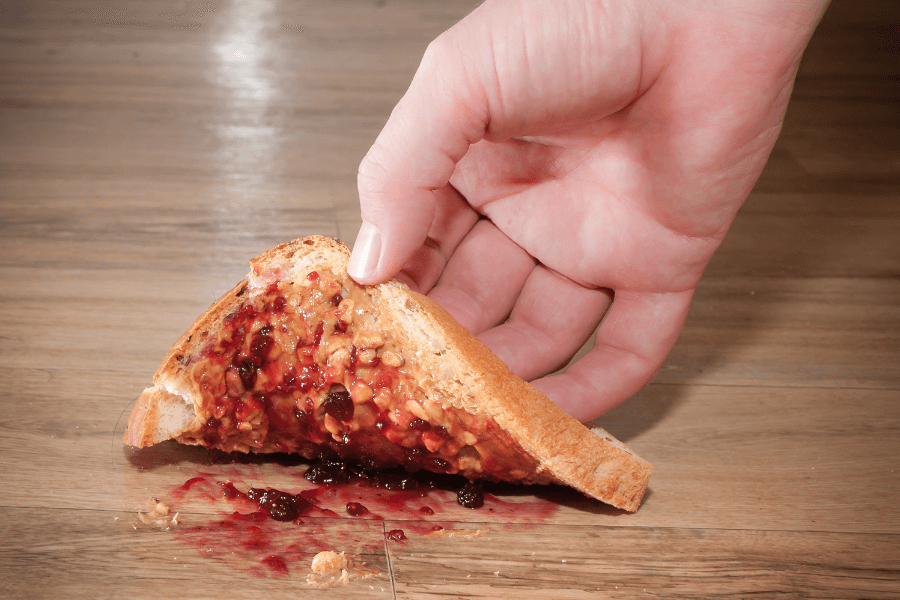 A close-up of a hand picking up a slice of peanut butter and jelly toast that has fallen face-down on a wooden floor. The jelly is smeared on the surface, creating a messy and unfortunate spill.