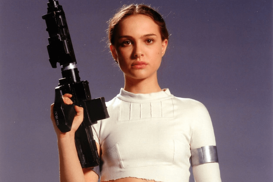 A woman with brown hair pulled back stands against a plain background, holding a large futuristic black blaster rifle. She wears a white, form-fitting cropped top with ribbed detailing, along with a silver armband on her upper left arm. Her expression is serious and determined.