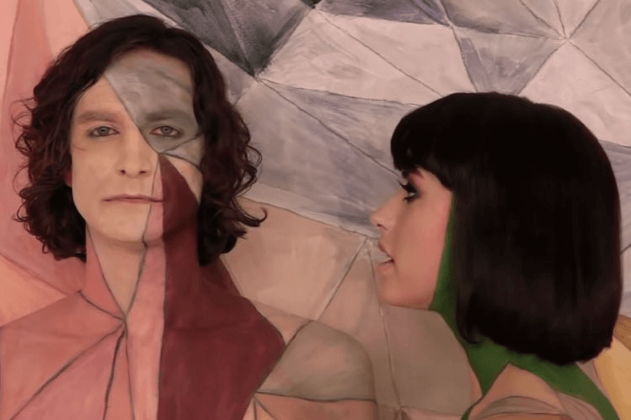 A man and a woman stand against a geometric, pastel-colored backdrop, their bodies painted to blend seamlessly with the background. The man faces forward with a solemn expression, while the woman, partially turned away, appears to be singing or speaking to him. The artistic body paint and stylized visuals suggest a scene from a music video or conceptual performance.