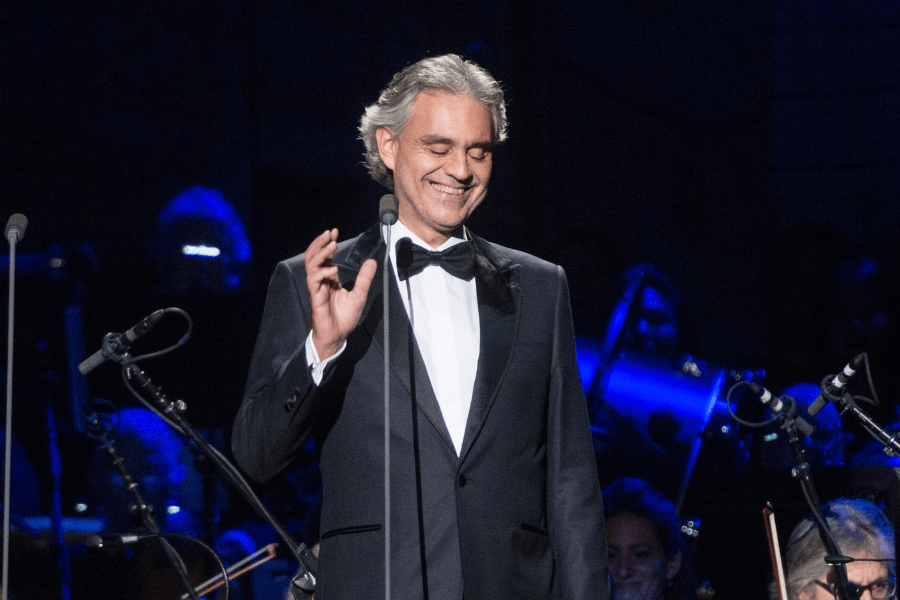 A male classical singer in a black tuxedo and bow tie stands on stage, smiling with his eyes closed while performing. He gestures gracefully with one hand near the microphone, exuding elegance and emotion. The dimly lit background features an orchestra with musicians illuminated by blue stage lighting.