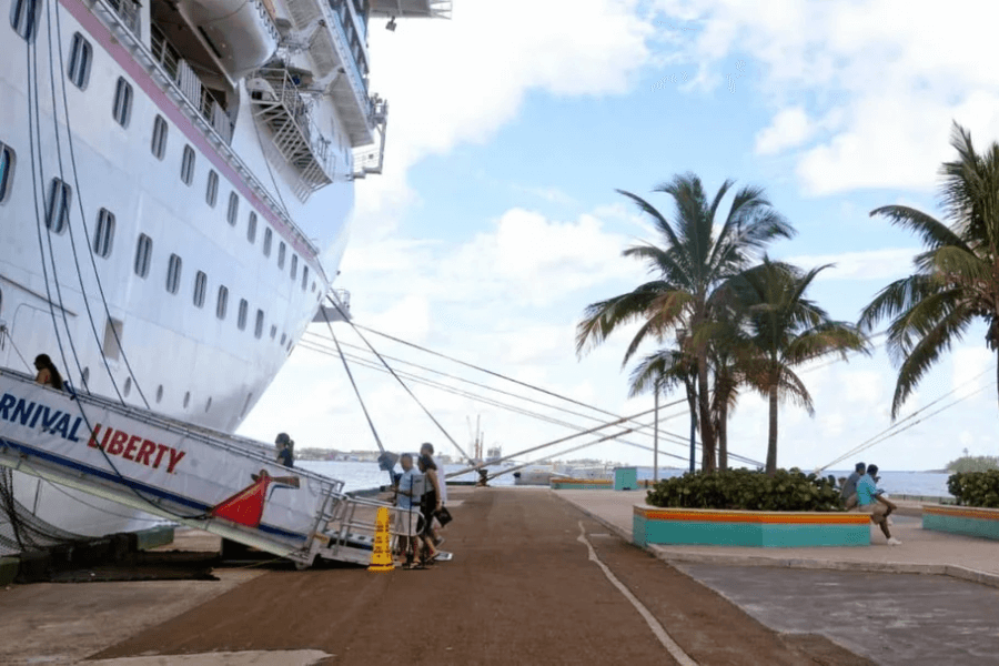 The Carnival Liberty cruise ship is docked at a tropical port, with passengers using the gangway to embark or disembark. Palm trees and a bright blue sky frame the pier, where a few people sit on benches enjoying the ocean view. Thick ropes secure the ship to the dock, emphasizing its massive size.