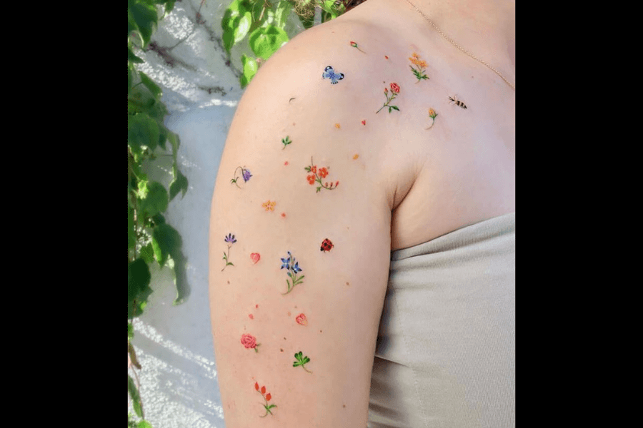 This image features a delicate tattoo of small, colorful flowers, hearts, and insects scattered across the upper arm and shoulder. The intricate designs include blue and orange flowers, a butterfly, a ladybug, and other nature-inspired details, arranged in a whimsical and organic pattern. The tattoo is vibrant and minimalistic, blending well with the natural tone of the skin.