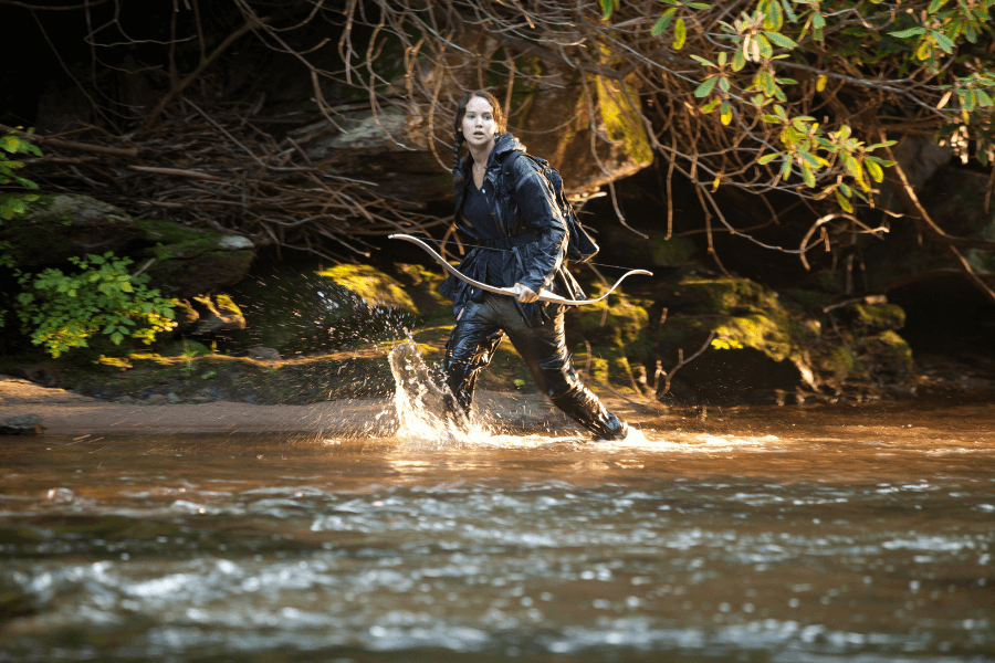 A young woman in a black survival outfit moves cautiously through a shallow river, gripping a bow. Her wet clothing and intense, focused expression suggest she is in a high-stakes situation, navigating the wilderness with urgency. The dense forest and rocky terrain in the background enhance the atmosphere of danger and survival.
