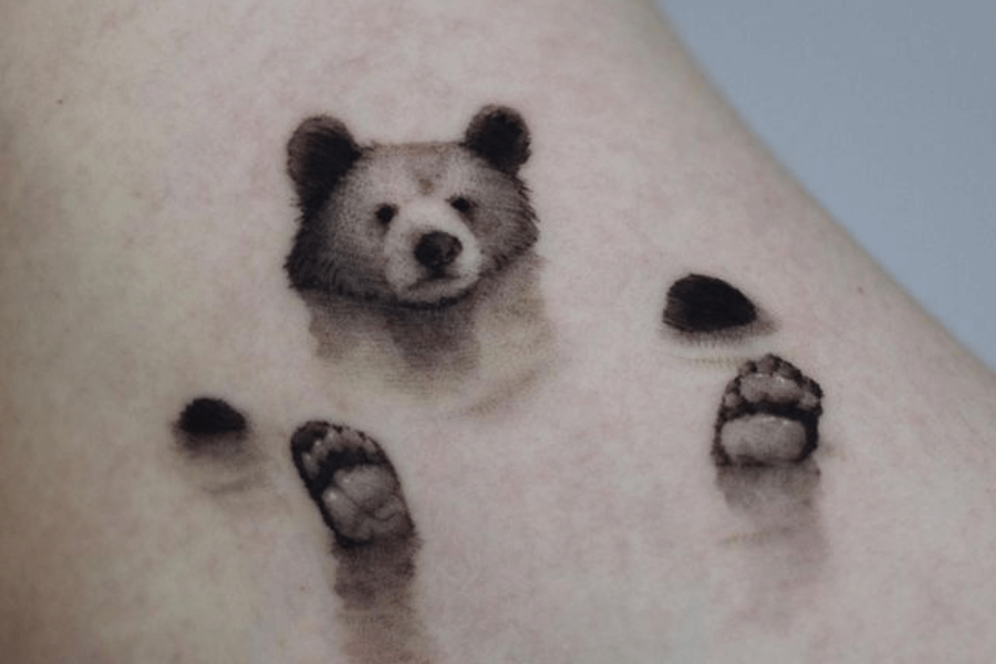 A tattoo of a small bear’s face with its two front paws below it, creating the illusion that the bear is playfully poking its head out of the skin. The design is soft and detailed, with a fuzzy, almost watercolor effect. The tattoo is minimalistic and placed on a light-toned patch of skin.
