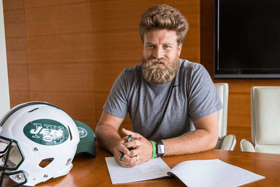 A bearded man with thick, wavy hair sits at a conference table, holding a pen and smiling slightly. He wears a gray t-shirt and sports a smartwatch and green wristband, with a New York Jets helmet and cap placed on the table beside him. The background features a wooden-paneled wall and a television screen, suggesting a formal signing or meeting.