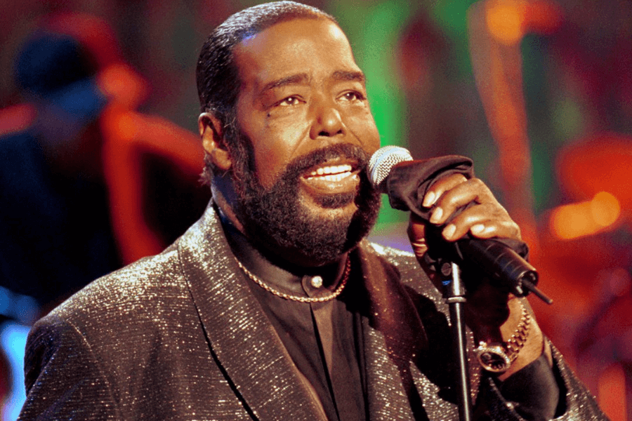 A male soul singer performs on stage, holding a microphone wrapped in a black cloth. He wears a shimmering black suit, a gold chain necklace, and a gold watch, exuding elegance and charisma. The warm stage lighting and blurred background of musicians enhance the intimate and emotional atmosphere of the performance.