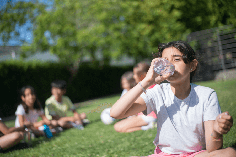 A young girl in a white t-shirt and pink shorts drinks water from a plastic bottle while sitting on the grass. In the blurred background, other children are sitting in a circle, likely taking a break during outdoor activities.