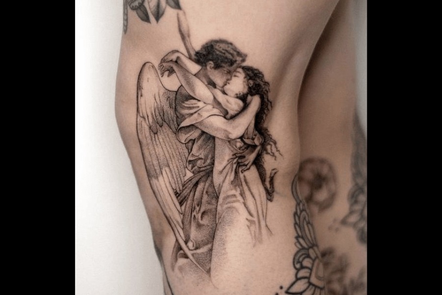 A highly detailed black and gray tattoo depicting an angel embracing a human figure. The angel has large, elegant wings, and both figures share an intimate and emotional connection in their pose. The artwork is shaded with soft, intricate lines, giving it a classical, almost Renaissance-style appearance.