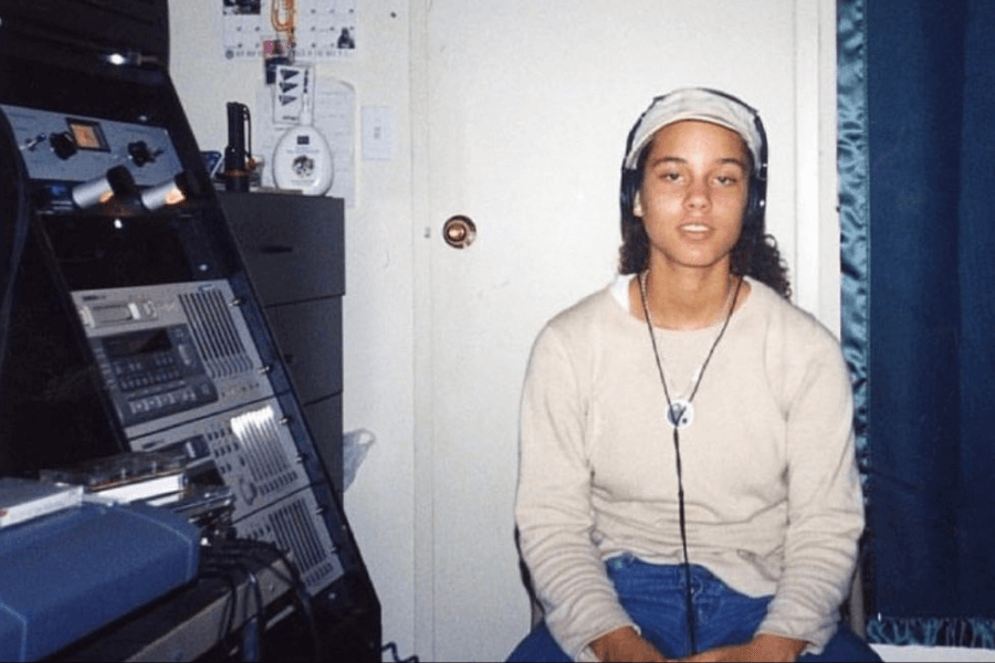 A young woman wearing a beige long-sleeve shirt, a headscarf, and headphones sits in a small recording or production room. She has a relaxed expression, with a peace sign pendant necklace around her neck. The background features a rack of audio equipment, a closed door, and a dark blue curtain, giving the space a home studio feel.