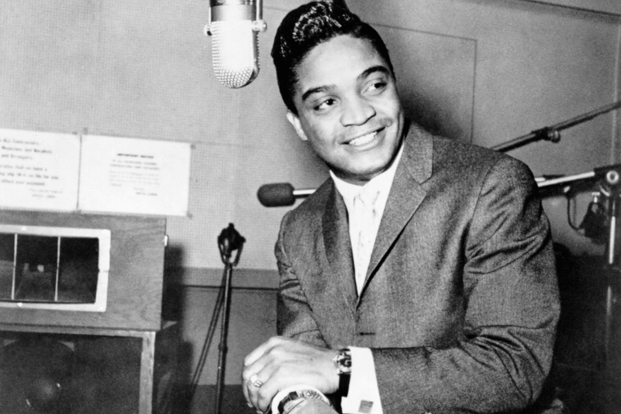 A black and white photograph of a male singer in a recording studio, smiling as he leans on a microphone. He is dressed in a sharp suit with a neatly styled pompadour, exuding charm and confidence. The vintage studio equipment and warm expression capture a timeless moment in music history.