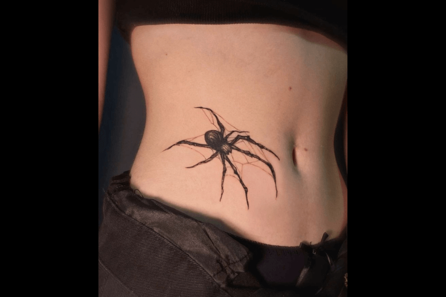 A tattoo of a large, detailed black spider positioned on the side of a person’s torso, near the waist. The spider has long, slender legs and intricate web-like details around it, giving the design a realistic and edgy appearance. The person is wearing black clothing, and the tattoo’s stark contrast with their skin makes it stand out.