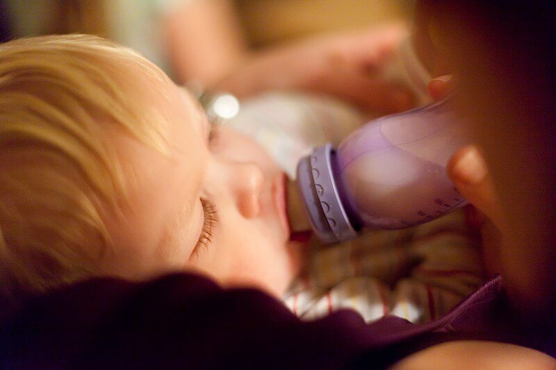 A close-up of a young child with blonde hair drinking from a baby bottle with a purple cap. The child is lying down, eyes partially closed, while an adult hand gently holds the bottle.