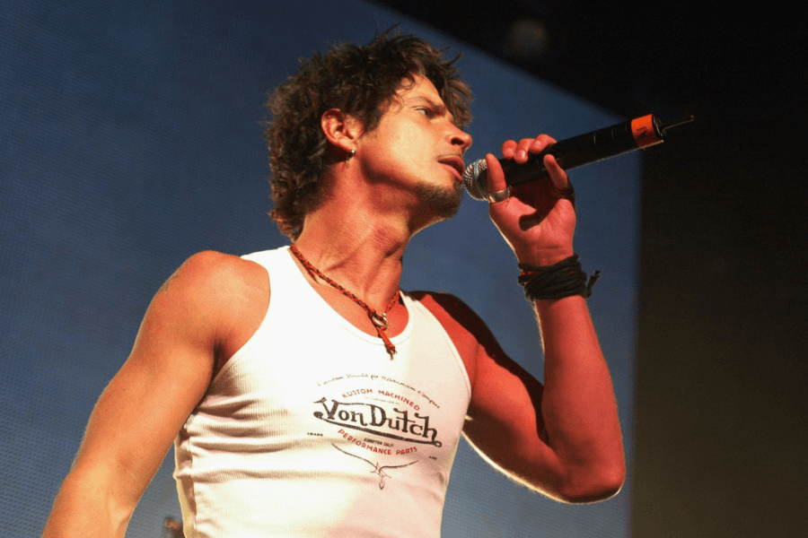 A male rock singer passionately performs on stage, gripping a microphone while tilting his head back. He wears a white Von Dutch tank top, a beaded necklace, and wristbands, exuding an intense and energetic presence. The stage lighting casts a warm glow, highlighting his expressive performance.