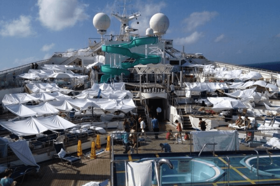 A cruise ship pool deck covered in makeshift white shade tents, likely improvised by passengers using bed sheets or towels. The deck is crowded with sun loungers, a water slide, and guests walking around or relaxing in the shade. The ship's upper deck and satellite domes are visible in the background under a bright blue sky.