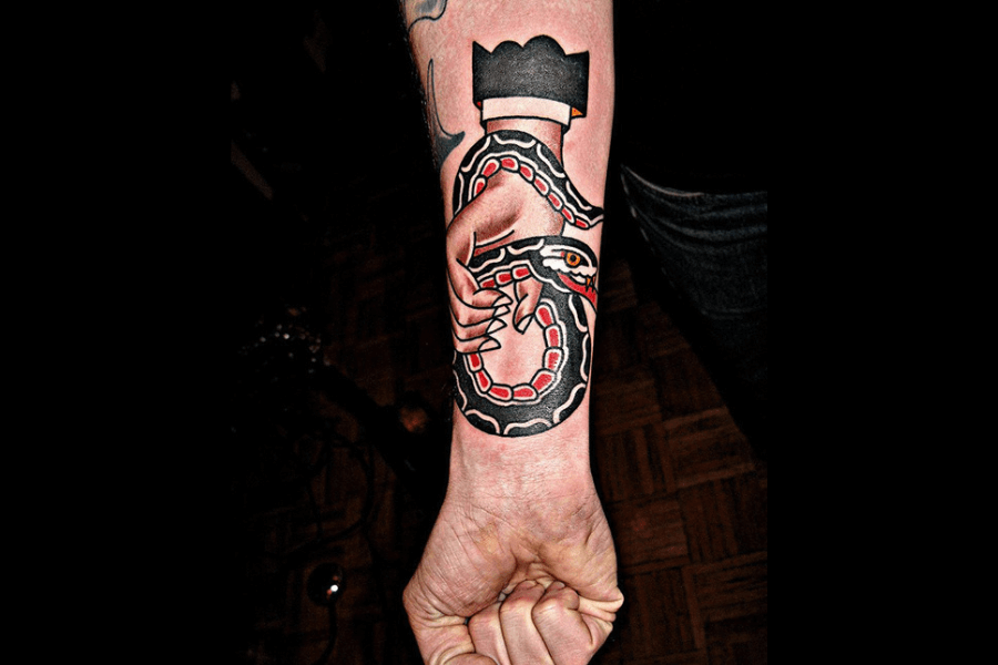 A bold, traditional-style tattoo featuring a hand gripping a coiled snake, with vivid red, black, and white patterns on the snake’s body. The hand appears strong, with sharp, dynamic lines, and a black crown hovers above the composition, giving the tattoo a striking and regal aesthetic. The design is placed on the forearm, showcasing clean, vibrant coloring and a classic tattoo vibe.