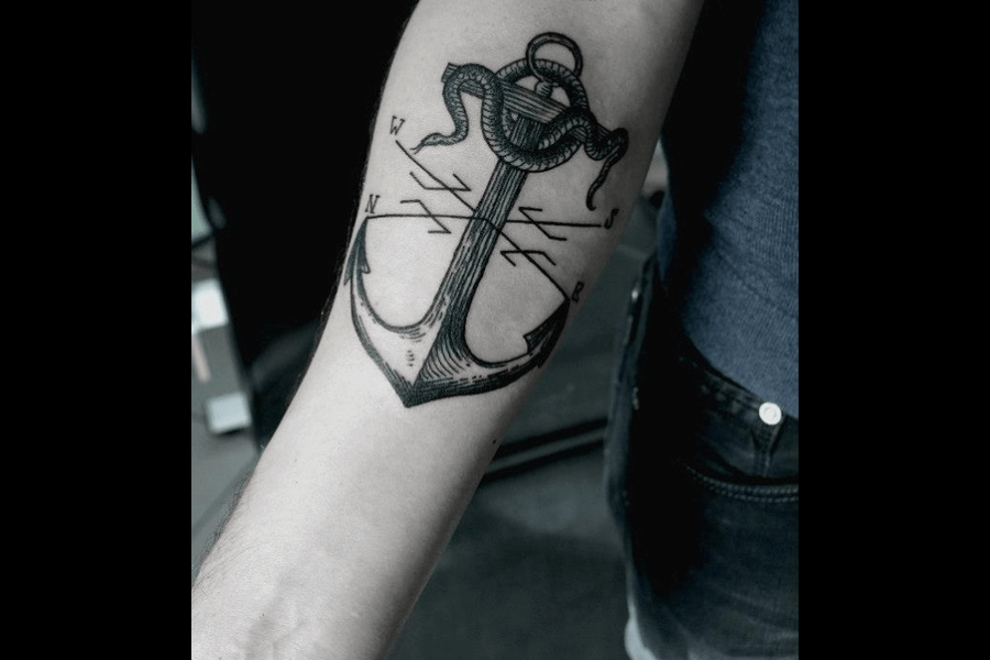 A black and gray tattoo featuring an anchor entwined with a snake, complemented by a compass design in the background. The anchor is detailed with fine linework, giving it a nautical yet modern feel, while the snake adds an element of danger and intrigue. The compass letters “N,” “S,” “E,” and “W” are prominently displayed, enhancing the theme of navigation and direction.