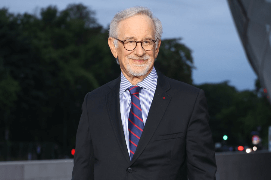 An older man with short white hair, glasses, and a trimmed beard smiles subtly while wearing a black suit, a blue striped shirt, and a red and blue striped tie. He stands outdoors in the evening, with a blurred background of trees, city lights, and modern architecture. His warm expression and formal attire give him a distinguished and professional appearance.