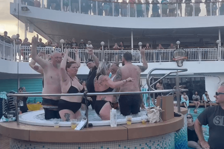  A lively scene at a cruise ship pool deck featuring a group of people dancing and raising their hands in a crowded hot tub. Many are in swimsuits, enjoying drinks while music plays. A crowd on the upper deck watches the fun, with some cheering and others relaxing by the pool.