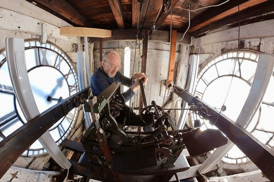 A clockmaker or maintenance worker is seen inside a large clock tower, adjusting the intricate mechanical gears of the clock’s mechanism. The aged interior features wooden beams, exposed wiring, and two large clock faces with Roman numerals glowing from the natural light outside. The worker, wearing glasses and a sweater, carefully handles the machinery, highlighting the craftsmanship involved in maintaining historic timepieces.