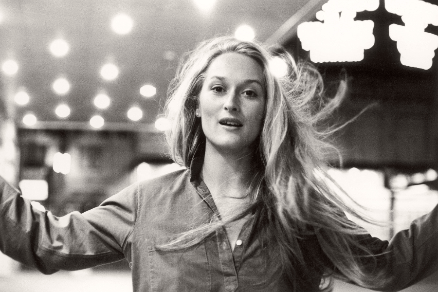 A young woman with long, flowing blonde hair gazes into the camera with a soft, confident expression. She wears a loose button-up shirt, and her arms are slightly extended, giving a sense of movement. The black-and-white setting, with blurred city lights in the background, creates a timeless and cinematic feel.