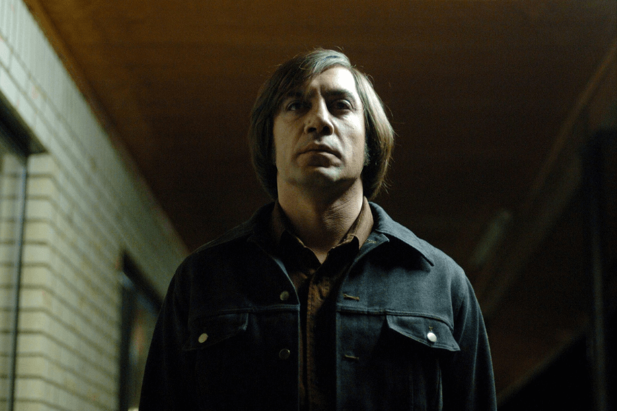 A man with a bowl-cut hairstyle and an emotionless expression stands in a dimly lit hallway. He wears a dark denim jacket over a brown shirt, his shadowed face adding to the eerie and unsettling atmosphere. The low lighting and tense framing create a sense of dread and quiet menace.