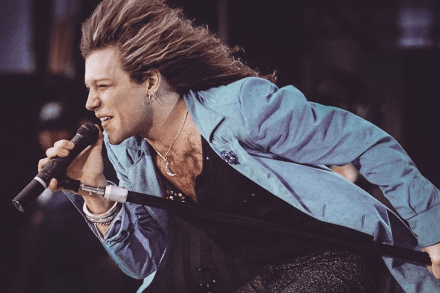 A male rock singer with long, flowing hair leans forward while gripping a microphone during an energetic stage performance. He wears a light blue blazer over a black, studded shirt, accessorized with silver bracelets, hoop earrings, and a crescent moon necklace. His intense expression and dynamic posture capture the electrifying spirit of the moment.