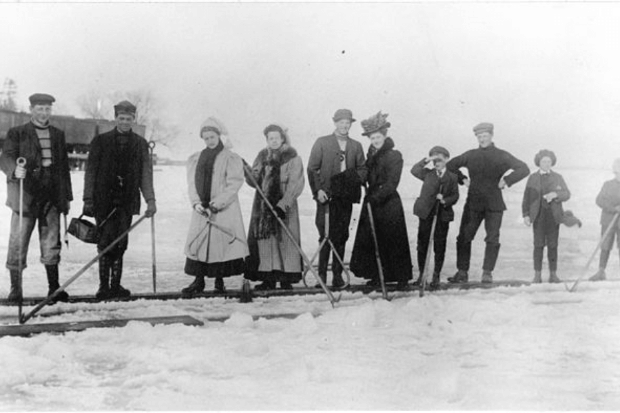 A historical black-and-white photograph of a group of men, women, and children standing on a frozen body of water, equipped with long sticks and dressed in early 20th-century winter attire. Some individuals wear heavy coats and hats, while others are in more casual work clothes, suggesting a mix of workers and recreational skaters. The group is gathered near an ice-cutting operation, possibly harvesting ice for storage or trade.