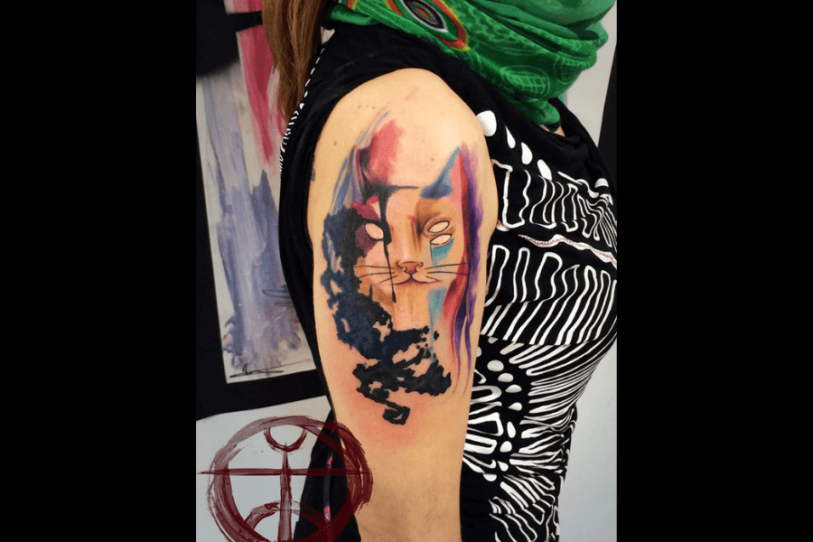 A colorful tattoo of a stylized cat on the upper arm, blending abstract and watercolor elements. The cat’s face is outlined with expressive, dynamic lines, featuring vibrant splashes of blue, purple, and pink, contrasted by bold black areas. The overall design has a contemporary, artistic feel, merging fluidity with sharp details.