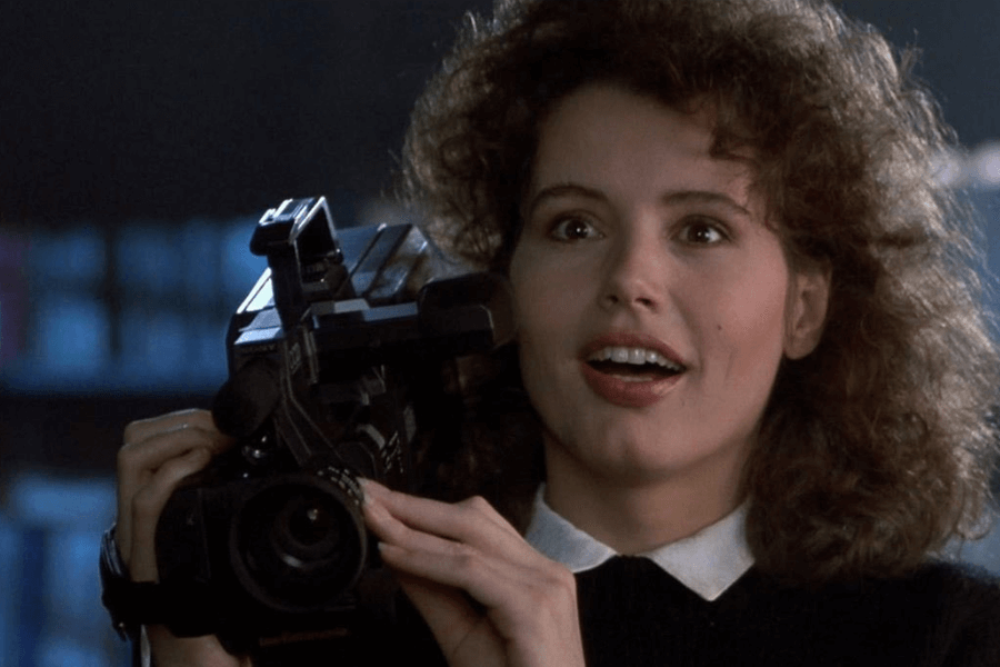 A woman with curly brown hair and an excited expression holds a vintage video camera up to her shoulder. She wears a black sweater over a white collared shirt, and her eyes are wide with anticipation as she looks at something off-camera. The dimly lit background suggests an indoor or nighttime setting, adding a cinematic feel to the moment.