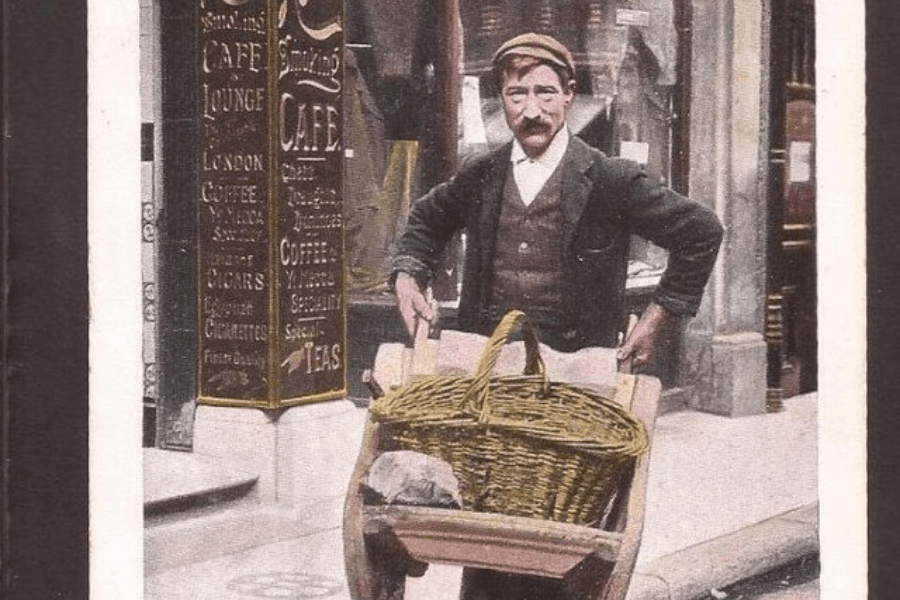 A vintage hand-colored photograph of a street vendor standing with a wooden handcart carrying a large wicker basket. The man, dressed in a cap, vest, and jacket, looks directly at the camera with a confident stance. Behind him, a café with ornate signage advertises coffee, cigars, and teas, evoking an early 20th-century urban setting.