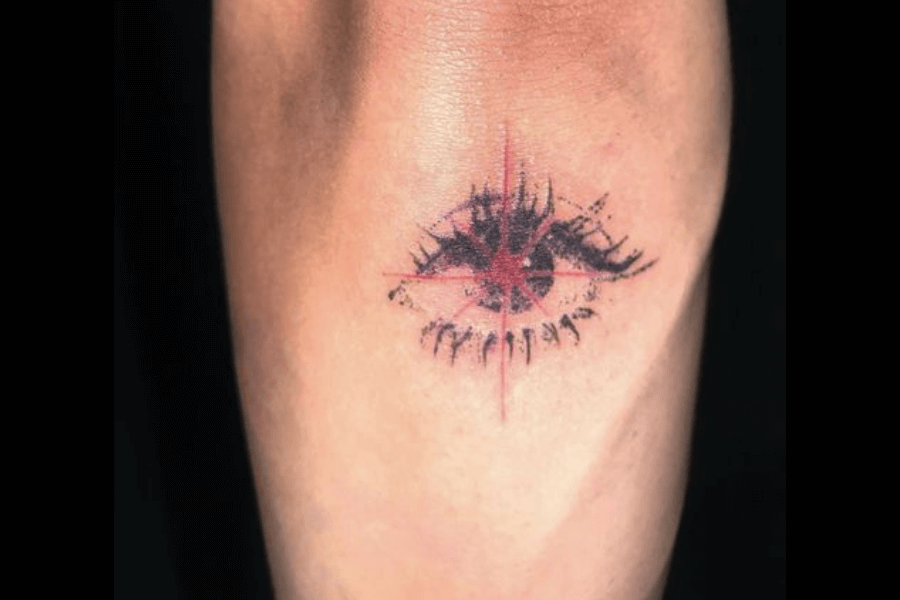 A minimalistic tattoo of an eye with dark, textured lashes and a striking red cross radiating through the center. The design combines abstract and symbolic elements, giving the eye a piercing, almost supernatural appearance. The shading and detailing create a sense of depth and mystery, with the red cross serving as a bold focal point.
