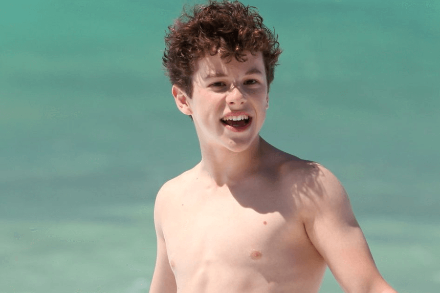 A young boy with curly brown hair smiles brightly while standing shirtless on a beach. The turquoise ocean water in the background glistens under the sunlight, adding to the carefree and joyful atmosphere. His expression conveys excitement and energy, as if enjoying a fun day by the sea.