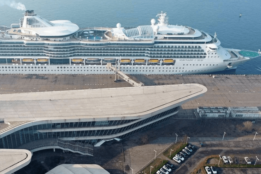  An aerial view of the Serenade of the Seas cruise ship docked at a modern port terminal. The ship's white exterior contrasts with the sleek, curved architecture of the terminal building, and a gangway connects the vessel to the pier. The calm water reflects the ship, while a parking lot with several cars is visible in the foreground.