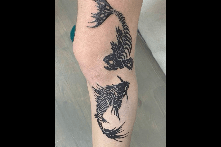 A tattoo of two fish skeletons in a yin-yang formation, done in bold black ink. The design emphasizes intricate details of the fish bones, with sharp, dramatic fins and tails, creating a striking and dynamic appearance. The overall composition suggests balance and duality, with an edgy, macabre twist.