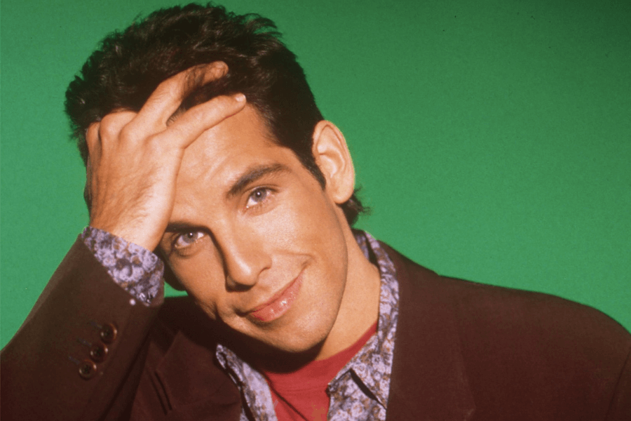 A man with dark hair and blue eyes smirks playfully while resting his hand on his forehead. He wears a burgundy blazer over a floral-patterned shirt with a red undershirt, set against a solid green background. His expression and pose convey a confident and humorous charm.