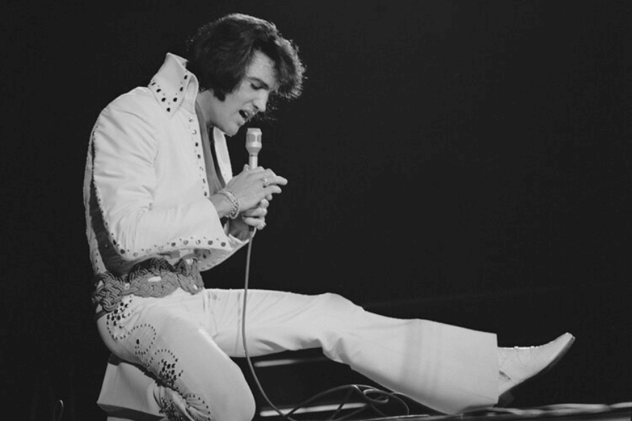 A black and white photograph of a male singer mid-performance, dressed in an embellished white jumpsuit with a high collar and a wide belt. He holds a microphone close to his mouth, singing with intensity while extending one leg outward in a dramatic pose. The dark background emphasizes his stage presence and iconic style.