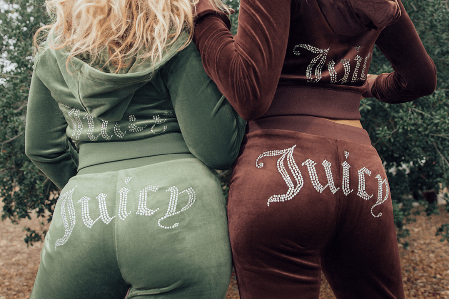 Two women wearing iconic Juicy Couture velour tracksuits, one in olive green and the other in rich brown, with rhinestone-embellished "Juicy" lettering across the back of their pants. The matching hoodies also feature sparkling text, adding to the glamorous early 2000s aesthetic. The nostalgic Y2K fashion look is captured from behind, emphasizing the signature bedazzled branding.