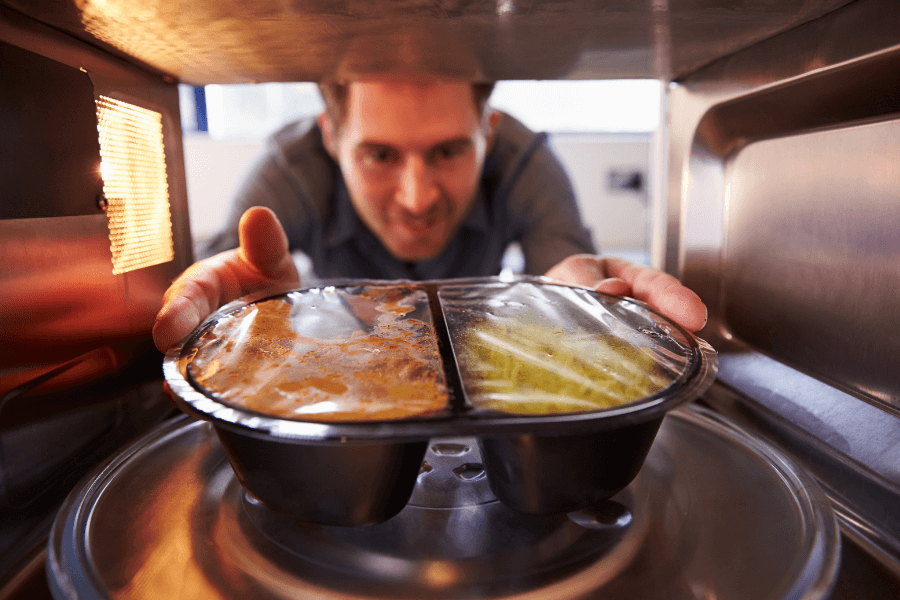 A man places a plastic-wrapped microwave meal inside a microwave oven, viewed from the inside. He has a focused and slightly eager expression as he prepares to heat his pre-packaged dinner.