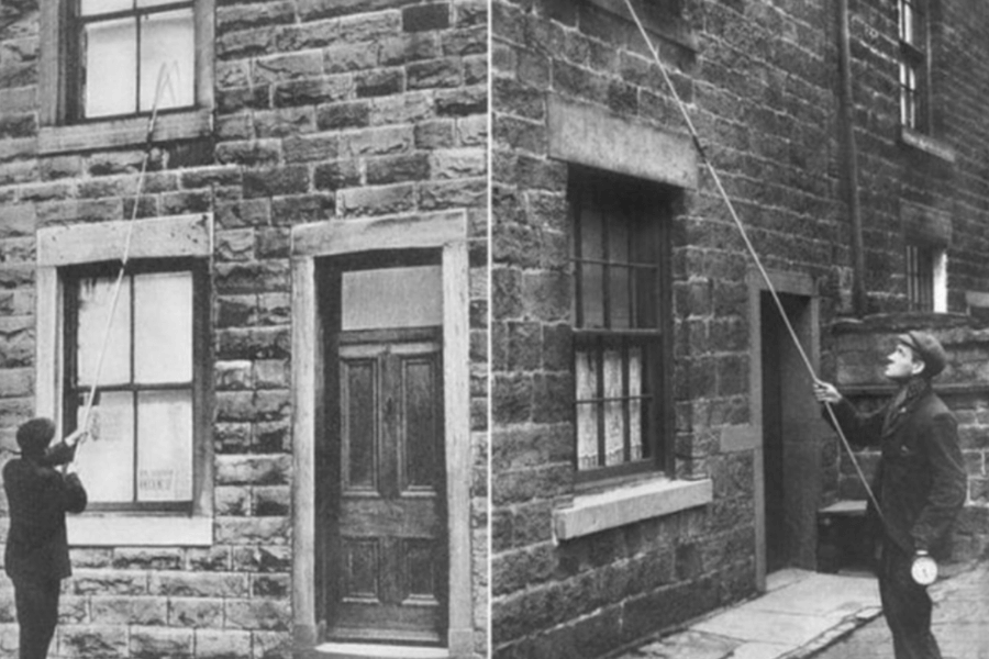 A black-and-white historical photograph showing two men standing outside stone-built houses, each using a long pole to tap on an upper-story window. The men are dressed in period-appropriate attire, including suits and caps, and appear to be performing the role of "knocker-uppers"—workers hired to wake people up before alarm clocks were common. One man holds a pocket watch, suggesting the importance of precise timing.