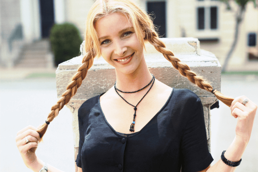 A smiling woman with long blonde hair styled into two braids playfully holds them out to the sides. She wears a black button-up top with short sleeves and layered black beaded necklaces. The background is slightly blurred, showing an outdoor urban setting with buildings and trees.