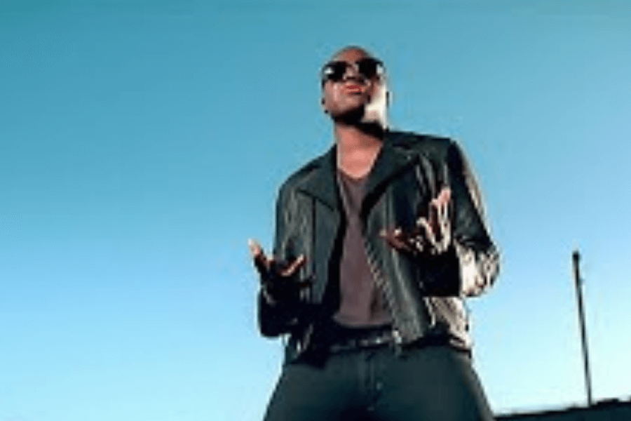 A man wearing a black leather jacket, dark sunglasses, and a black shirt stands against a clear blue sky, gesturing expressively with his hands. His stance and facial expression convey emotion, possibly mid-performance or in a dramatic music video scene. The lighting and framing emphasize a cool, cinematic aesthetic.