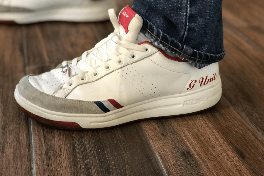 A close-up of a person wearing classic white and red Reebok G-Unit sneakers, a signature shoe collaboration from the early 2000s. The sneaker features a clean leather upper with perforated detailing, a suede toe cap, red and blue striped accents, and the "G-Unit" branding embroidered on the side. The design reflects the hip-hop and streetwear influence of the era, made popular by 50 Cent and G-Unit.