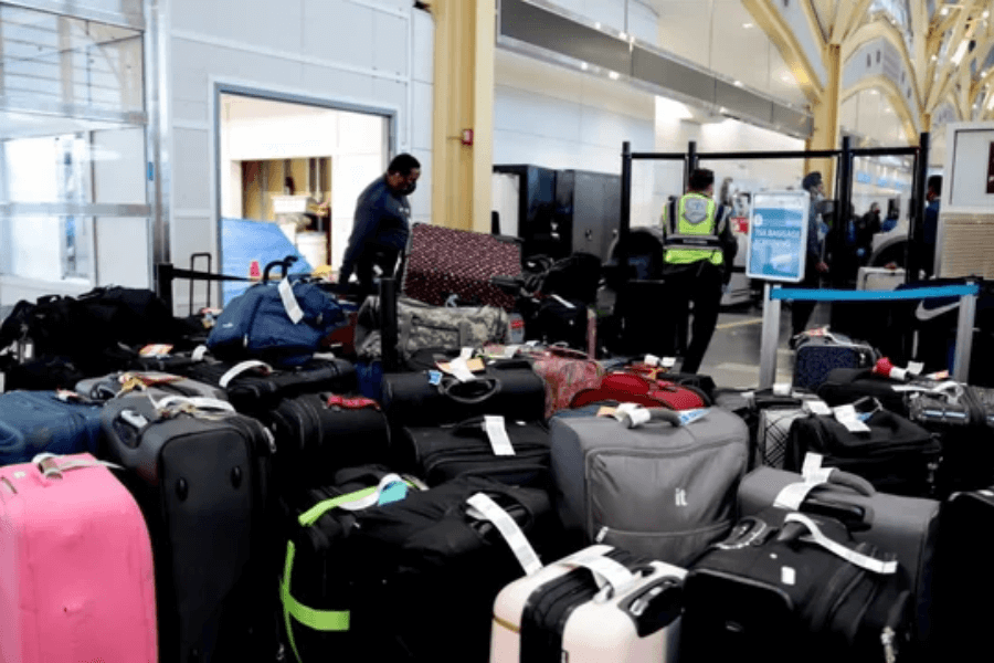 A crowded baggage claim area filled with suitcases of various colors, sizes, and styles, many with luggage tags attached. Airport or cruise port staff in uniforms are seen handling the bags, while a security officer stands near a checkpoint. The scene suggests a busy travel hub with passengers waiting to collect their belongings.