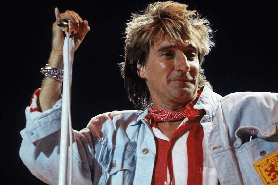 A male rock singer performs on stage, holding onto a microphone stand wrapped in a white scarf. He wears a red and white striped shirt, a light-wash denim jacket, and a red polka-dotted scarf around his neck. His tousled blond hair and confident smirk capture the energetic and charismatic essence of his performance.