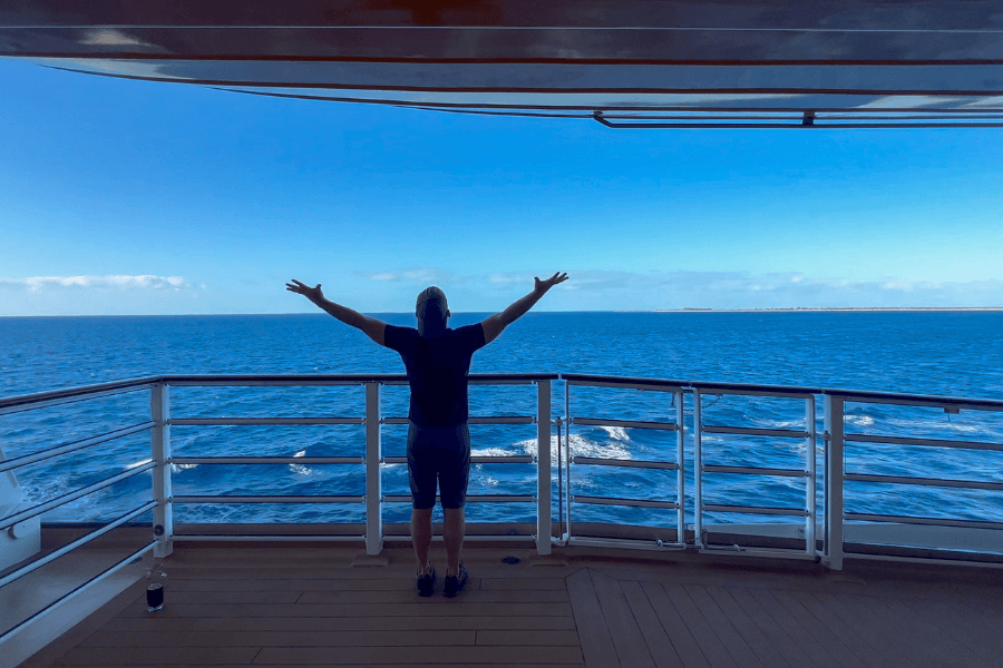 A person stands at the railing of a cruise ship's outdoor deck with arms outstretched, embracing the vast ocean view. The deep blue water stretches to the horizon, with a distant landmass faintly visible under a clear sky. The peaceful moment captures the essence of freedom and relaxation at sea.