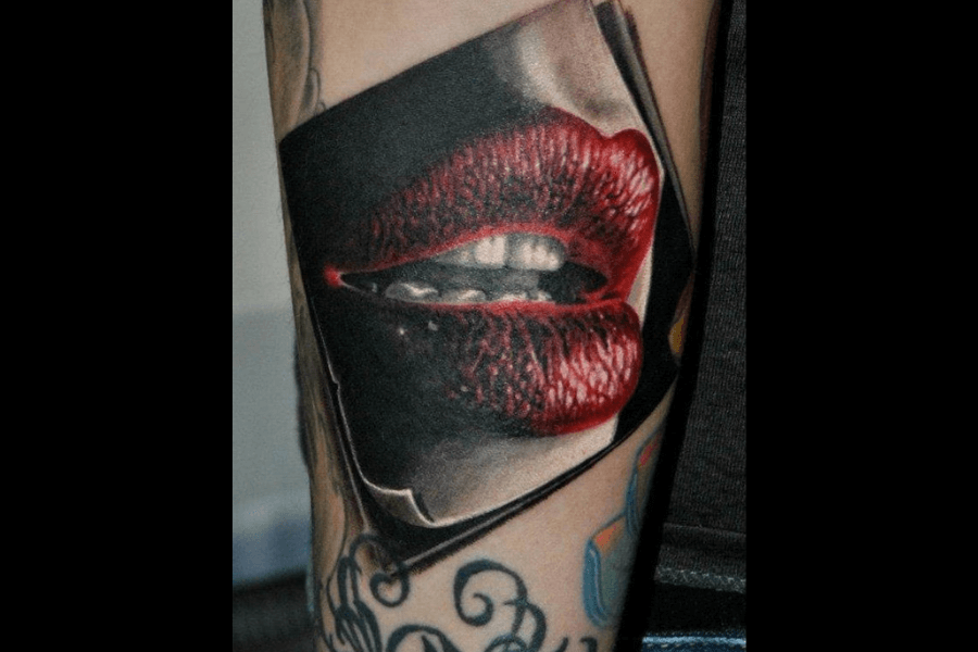 A hyper-realistic tattoo of red, glossy lips slightly parted to reveal teeth, with intricate detail emphasizing the texture and shine. The lips are framed within a bold, rectangular border, creating a striking visual focus. Surrounding the design are faint hints of other tattoos on the arm, adding depth to the composition.
