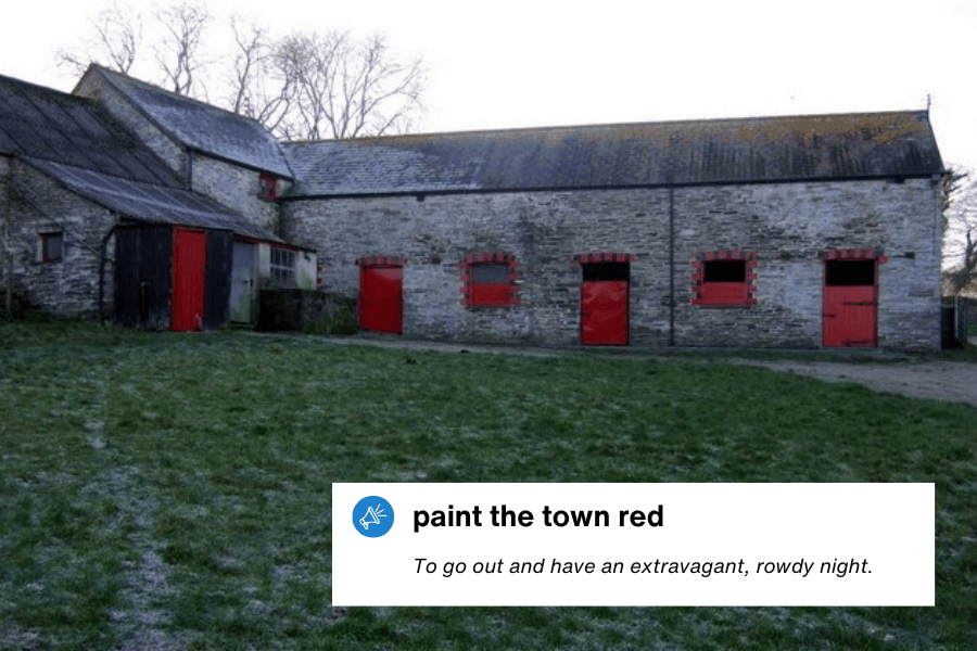 A rustic stone building with red-painted doors and window frames stands against a frosty ground, giving a striking contrast to the otherwise muted surroundings. The weathered exterior suggests an old barn or stable. A text overlay in the bottom left corner reads: "Paint the town red – To go out and have an extravagant, rowdy night."