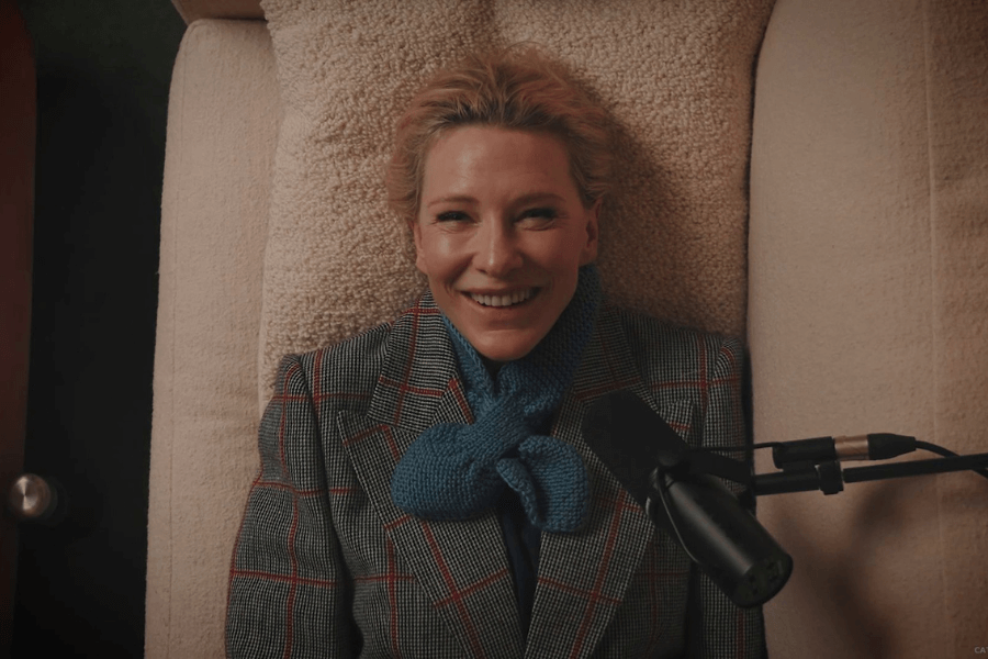 A woman with short blonde hair lies on a plush beige couch, smiling warmly at the camera. She wears a checkered blazer with red accents and a cozy blue knitted scarf, exuding a relaxed yet stylish look. A microphone is positioned near her, suggesting a podcast or interview setting.