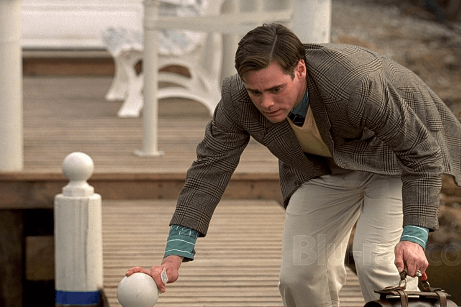 A man in a plaid blazer and beige pants leans down on a wooden dock, touching a white railing post that appears artificial. He has a curious and slightly suspicious expression, holding a leather briefcase in his other hand. The scene suggests a moment of realization or discovery in a seemingly perfect but controlled environment.